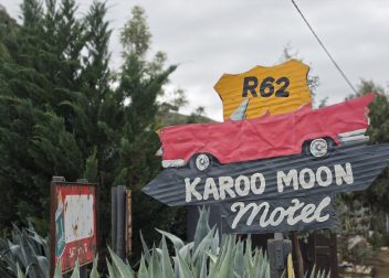 Rustic Route 62 sign for Karoo Moon Motel, featuring a red vintage car, surrounded by agave plants and trees.