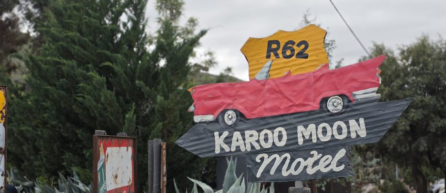 Rustic Route 62 sign for Karoo Moon Motel, featuring a red vintage car, surrounded by agave plants and trees.